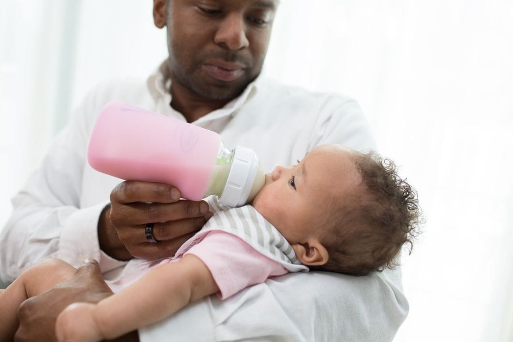 Vader houdt zijn baby in de armen terwijl hij melk geeft met een Dr Brown brede halsfles met roze beschermhoes, een praktische dr brown babyfles verkrijgbaar bij Happy Kids Boutique met aandacht voor comfort en duurzaamheid.