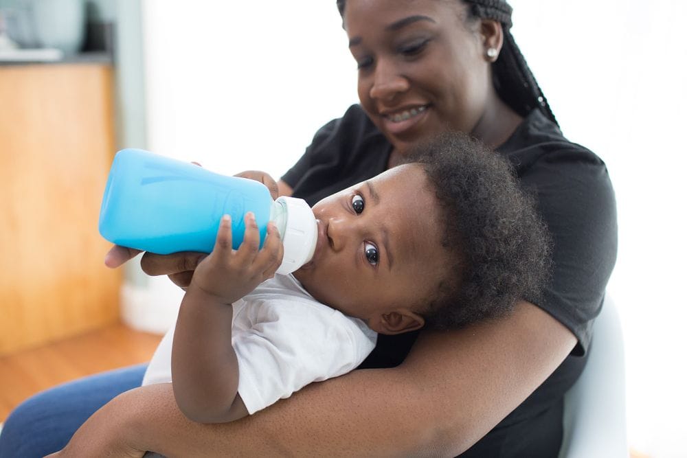Moeder houdt haar kind op schoot terwijl hij drinkt uit een Dr Brown brede halsfles met blauwe beschermhoes, een comfortabele voedingssituatie met een dr brown babyfles verkrijgbaar bij Happy Kids Boutique met aandacht voor duurzaamheid.