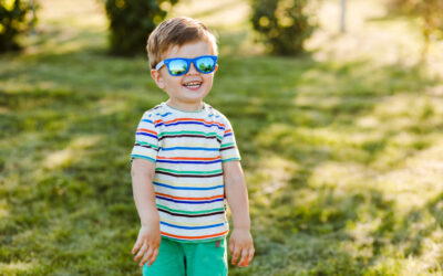 Happy Kids Boutique kind staat lachend in het gras en draagt een blauwe zonnebril, vrolijke baby met gestreept shirt geniet van het buitenspelen in een zonnige omgeving