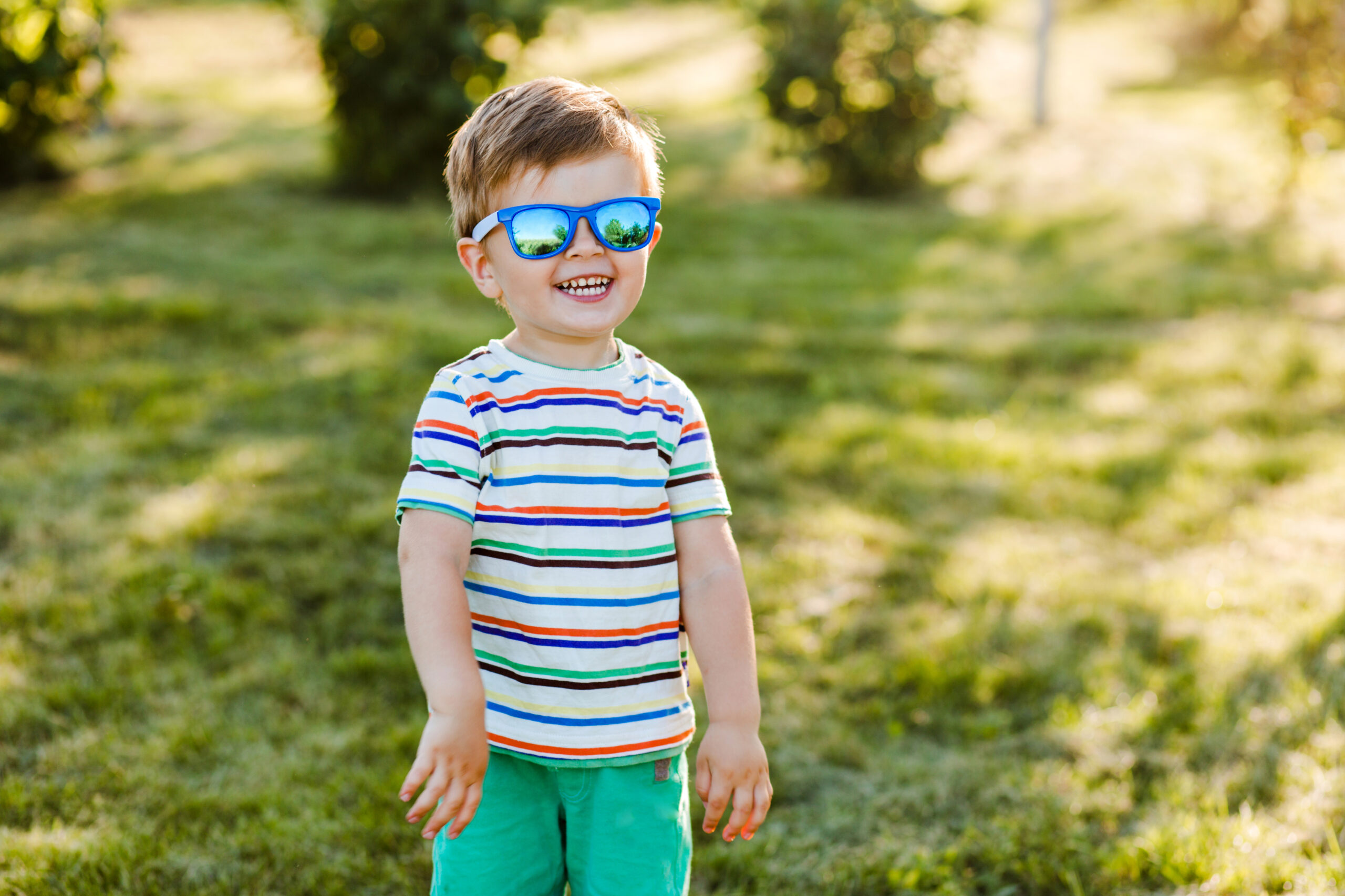 Blogfoto babyzonnebril kopen Happy Kids Boutique kind staat lachend in het gras en draagt een blauwe zonnebril, vrolijke baby met gestreept shirt geniet van het buitenspelen in een zonnige omgeving