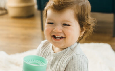 Een lachende baby zit op een zacht wit kleed in een warme woonkamer en houdt een groene drinkbeker vast met vrolijke print. De drinkbeker van Dr. Brown, ook bekend als dr brown 360° drinkbeker, ligt fijn in kleine handjes en ondersteunt zelfstandig drinken. Deze sfeervolle afbeelding past bij het assortiment van Happy Kids Boutique en laat zien hoe een praktische drinkbeker bijdraagt aan fijne dagelijkse momenten voor jonge gezinnen.