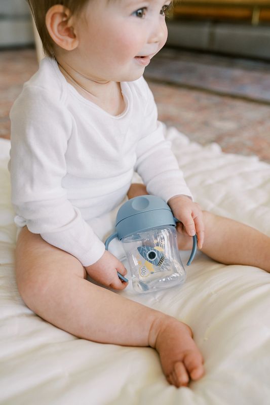Een baby zit op een zacht bed en houdt een blauwe drinkbeker met handvatten vast, terwijl hij nieuwsgierig vooruit kijkt. De beker is de Dr. Brown Trainingsbeker zachte tuit 180 ml, ook bekend als dr brown trainingsbeker, met transparante cup en speelse print. Deze Trainingsbeker zachte tuit 180 ml is verkrijgbaar bij Happy Kids Boutique en ondersteunt de overgang naar zelfstandig drinken op een comfortabele manier.