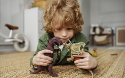 Kind speelt geconcentreerd op de vloer met twee dinosaurusspeelgoed figuren en bedenkt een fantasierijk spelmoment. Op de achtergrond is een lichte kinderkamer zichtbaar. Deze sfeerafbeelding van Happy Kids Boutique laat zien hoe spelen belangrijk is voor de ontwikkeling van kinderen met aandacht voor creativiteit en duurzaamheid.
