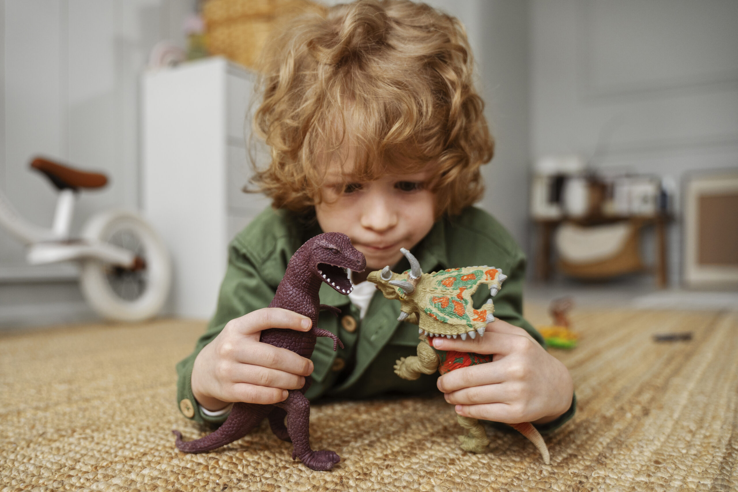 Kind speelt geconcentreerd op de vloer met twee dinosaurusspeelgoed figuren en bedenkt een fantasierijk spelmoment. Op de achtergrond is een lichte kinderkamer zichtbaar. Deze sfeerafbeelding van Happy Kids Boutique laat zien hoe spelen belangrijk is voor de ontwikkeling van kinderen met aandacht voor creativiteit en duurzaamheid.