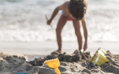 Kind speelt op het strand tijdens een zonnige stranddag en graaft in het zand met speelgoed zoals een schepje en harkje, terwijl de zee op de achtergrond zichtbaar is. Deze sfeerafbeelding van Happy Kids Boutique laat zien hoe kinderen onbezorgd spelen en ontdekken met aandacht voor een bewuste levensstijl en duurzaamheid.