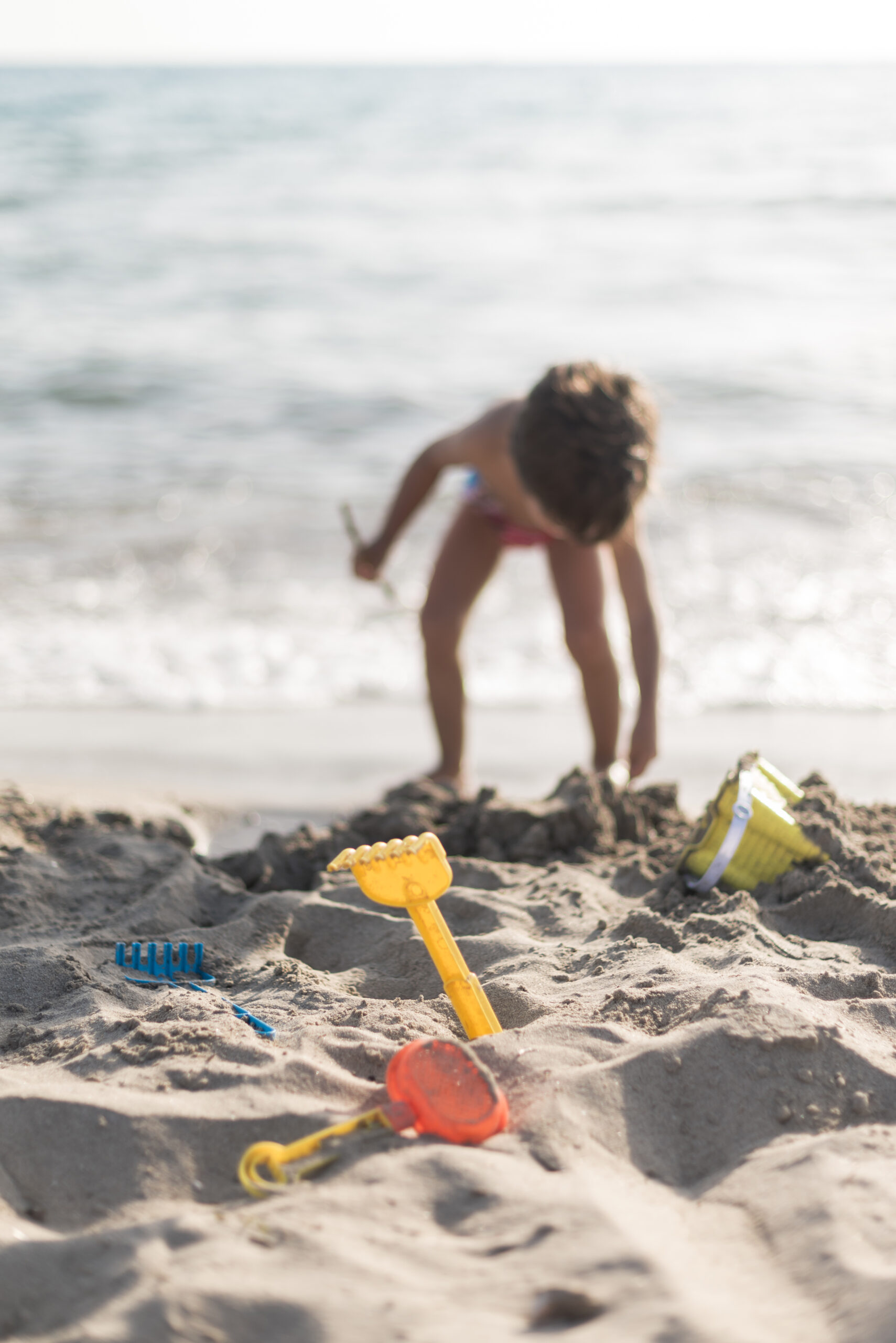 Kind speelt op het strand tijdens een zonnige stranddag en graaft in het zand met speelgoed zoals een schepje en harkje, terwijl de zee op de achtergrond zichtbaar is. Deze sfeerafbeelding van Happy Kids Boutique laat zien hoe kinderen onbezorgd spelen en ontdekken met aandacht voor een bewuste levensstijl en duurzaamheid.