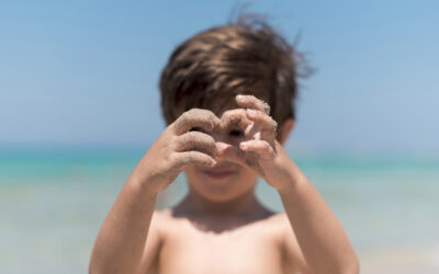 Happy Kids Boutique sfeerbeeld van een kind dat op het strand met zanderige handen speelt en zand vasthoudt voor zijn gezicht met de zee op de achtergrond, passend bij natuurlijk buitenspelen en duurzaamheid
