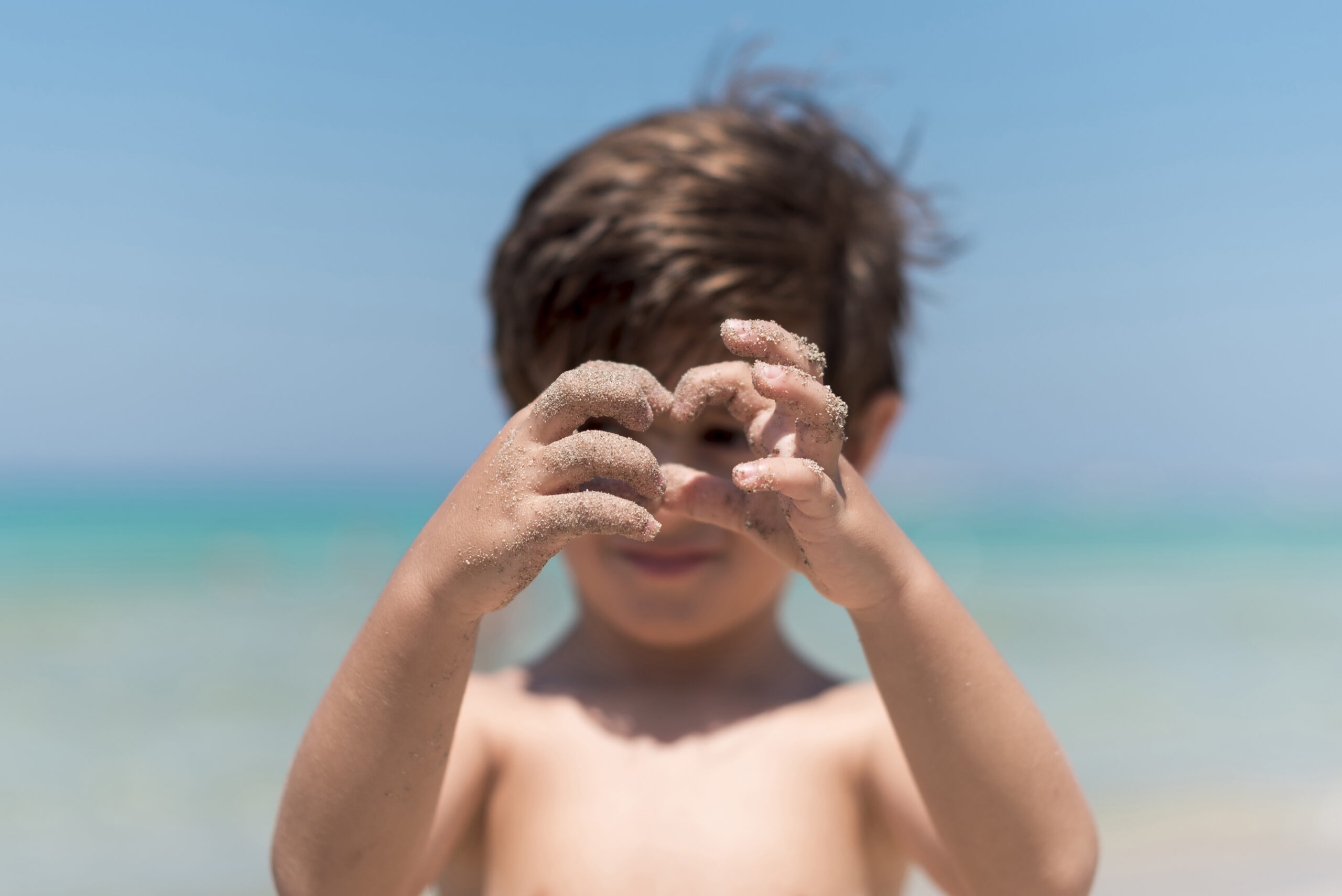 Blogfoto strand hacks Happy Kids Boutique sfeerbeeld van een kind dat op het strand met zanderige handen speelt en zand vasthoudt voor zijn gezicht met de zee op de achtergrond, passend bij natuurlijk buitenspelen en duurzaamheid