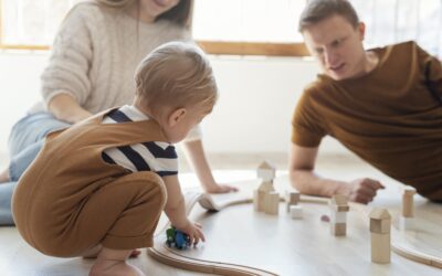 Kind speelt met een houten treinbaan op de vloer terwijl ouders meespelen en betrokken toekijken in een lichte woonkamer. De houten blokken en trein zorgen voor een rustig spelmoment samen. Deze sfeerafbeelding van Happy Kids Boutique laat zien hoe spelen met kinderen en ouders meespelen bijdraagt aan verbinding en ontwikkeling met aandacht voor duurzaamheid.
