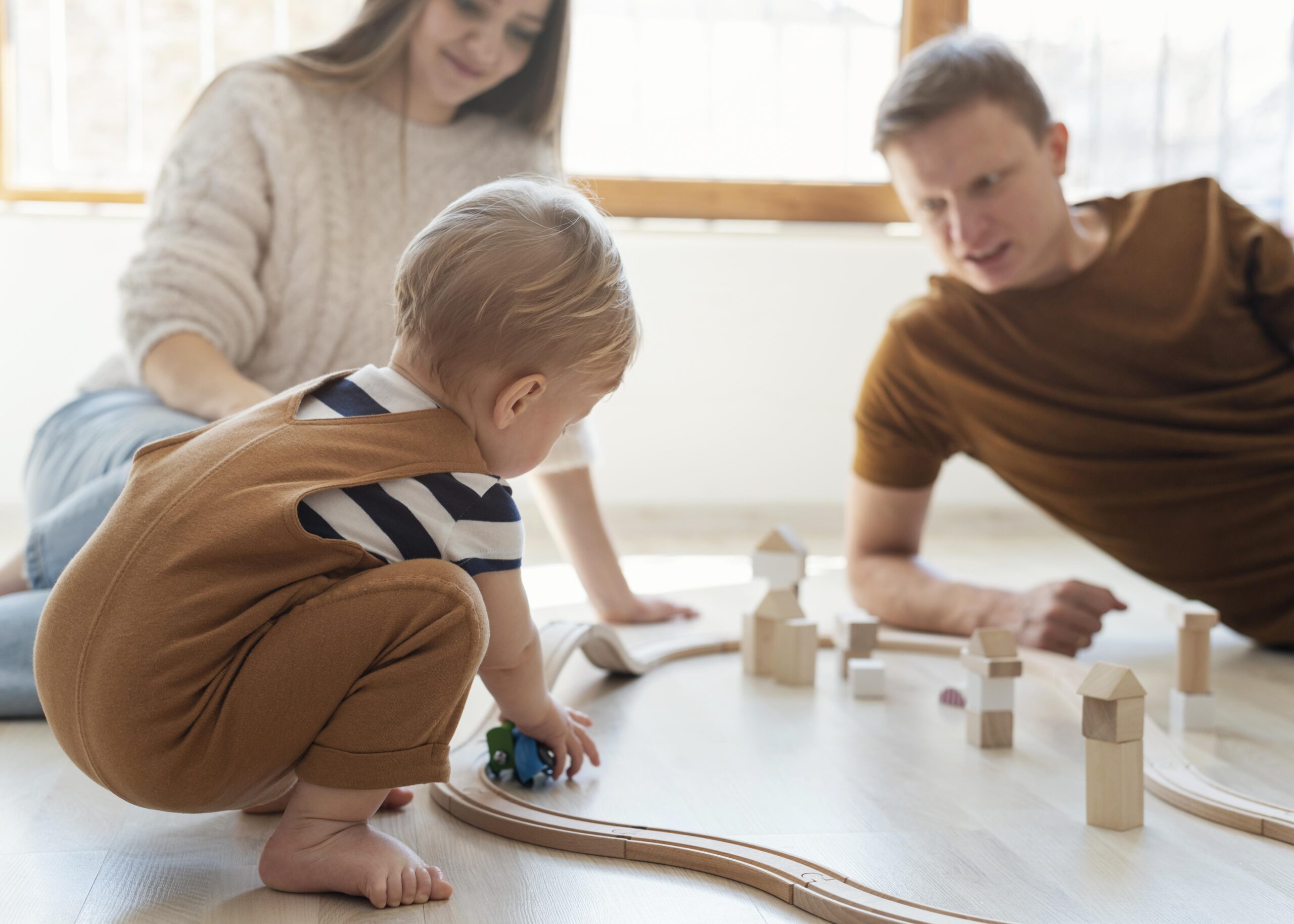 Kind speelt met een houten treinbaan op de vloer terwijl ouders meespelen en betrokken toekijken in een lichte woonkamer. De houten blokken en trein zorgen voor een rustig spelmoment samen. Deze sfeerafbeelding van Happy Kids Boutique laat zien hoe spelen met kinderen en ouders meespelen bijdraagt aan verbinding en ontwikkeling met aandacht voor duurzaamheid.