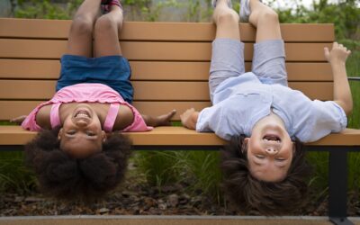 Happy Kids Boutique diversiteit en cultuurverschillen zichtbaar in twee kinderen die lachend ondersteboven op een bankje buiten hangen en samen spelen in een natuurlijke omgeving met aandacht voor duurzaamheid