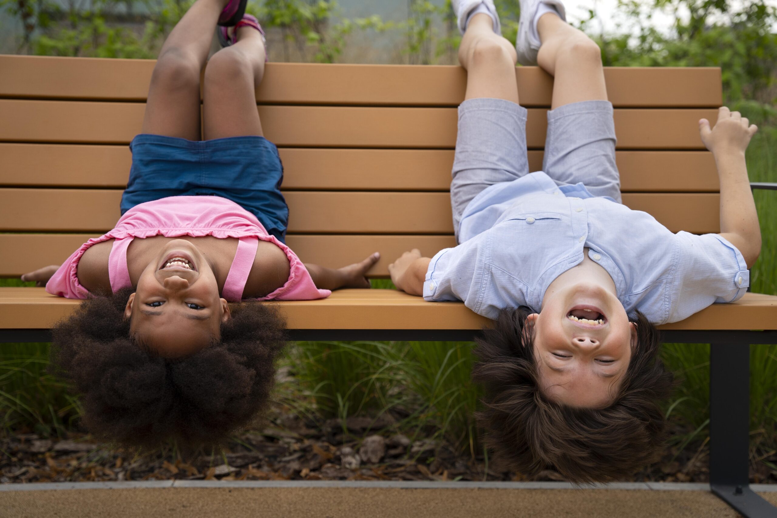 Happy Kids Boutique diversiteit en cultuurverschillen zichtbaar in twee kinderen die lachend ondersteboven op een bankje buiten hangen en samen spelen in een natuurlijke omgeving met aandacht voor duurzaamheid