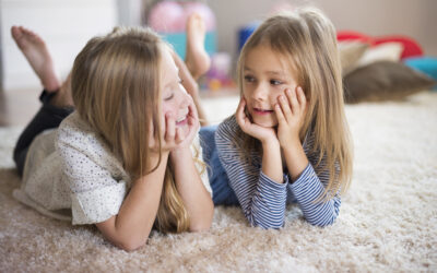 Twee kinderen liggen samen op een zacht vloerkleed en kijken elkaar lachend aan in een lichte woonkamer, een warm en speels moment dat past bij Happy Kids Boutique waar babykleding en babybenodigdheden samenkomen met aandacht voor comfort en duurzaamheid. Afbeelding designes by Freepik.