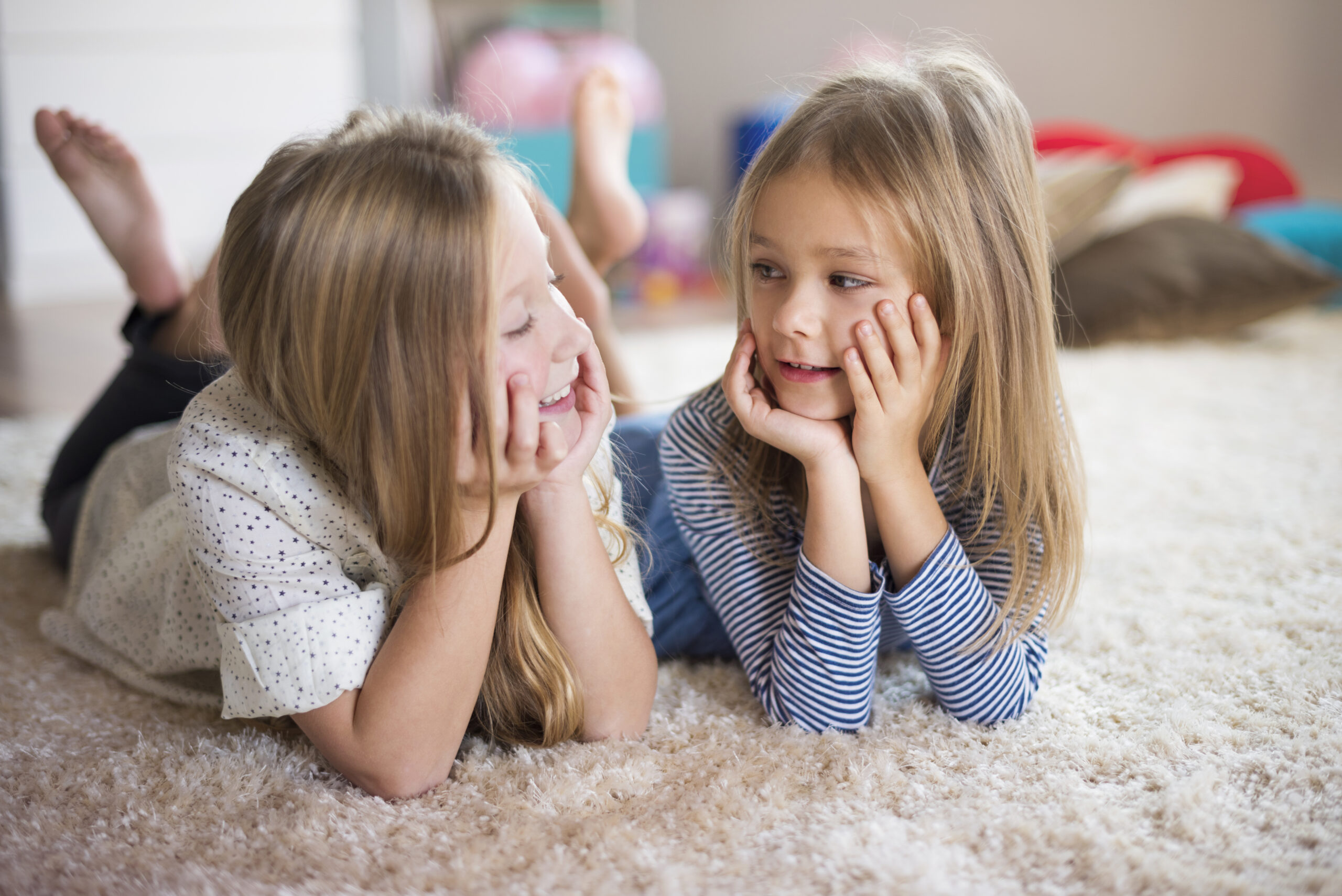 Twee kinderen liggen samen op een zacht vloerkleed en kijken elkaar lachend aan in een lichte woonkamer, een warm en speels moment dat past bij Happy Kids Boutique waar babykleding en babybenodigdheden samenkomen met aandacht voor comfort en duurzaamheid. Afbeelding designes by Freepik.