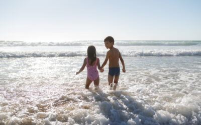 Twee kinderen lopen hand in hand de zee in op het strand in Nederland terwijl de golven om hen heen bewegen en de zon laag aan de horizon staat. Deze warme sfeerafbeelding van Happy Kids Boutique laat zien hoe kinderen samen spelen en genieten van een strandmoment met aandacht voor een bewuste levensstijl en duurzaamheid.