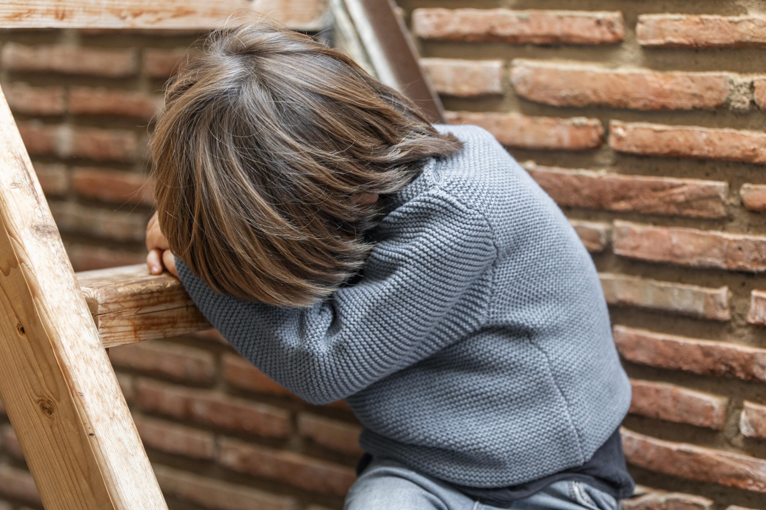 Happy Kids Boutique kind zit buiten op een houten trap met hoofd op de armen en oogt boos en gefrustreerd tegen een bakstenen muur, moment van emotie met aandacht voor duurzaamheid