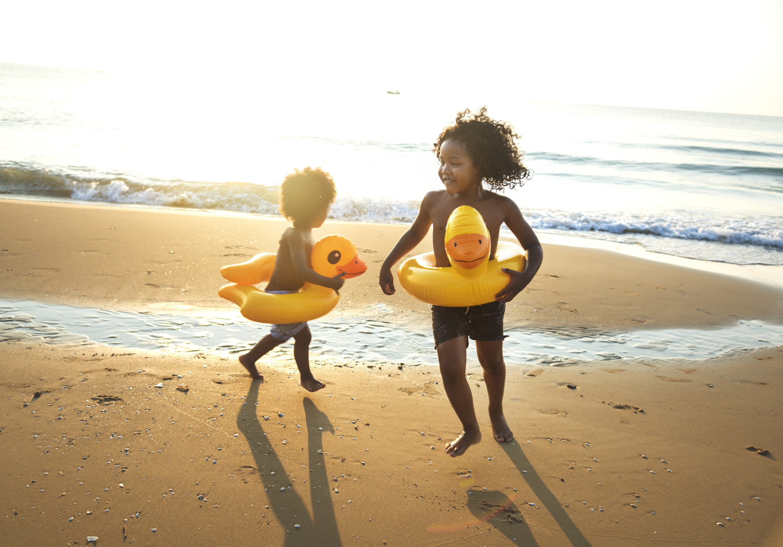 Twee kinderen spelen en rennen op het strand tijdens een zonnige strandvakantie met opblaasbare zwemringen in de vorm van eenden. De zee en het zand vormen een warme achtergrond terwijl de kinderen plezier maken aan de waterlijn. Deze sfeerafbeelding van Happy Kids Boutique straalt vrijheid, spelen en samenzijn uit met aandacht voor een bewuste en duurzame levensstijl en duurzaamheid.