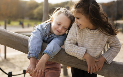 Twee kinderen leunen samen op een houten speeltoestel en lachen terwijl ze dicht tegen elkaar aan zitten in een buitenomgeving. Het moment straalt warmte en vriendschap uit tussen de kinderen tijdens het spelen. Deze sfeerafbeelding van Happy Kids Boutique benadrukt verbinding, samenzijn en een bewuste levensstijl met aandacht voor duurzaamheid.