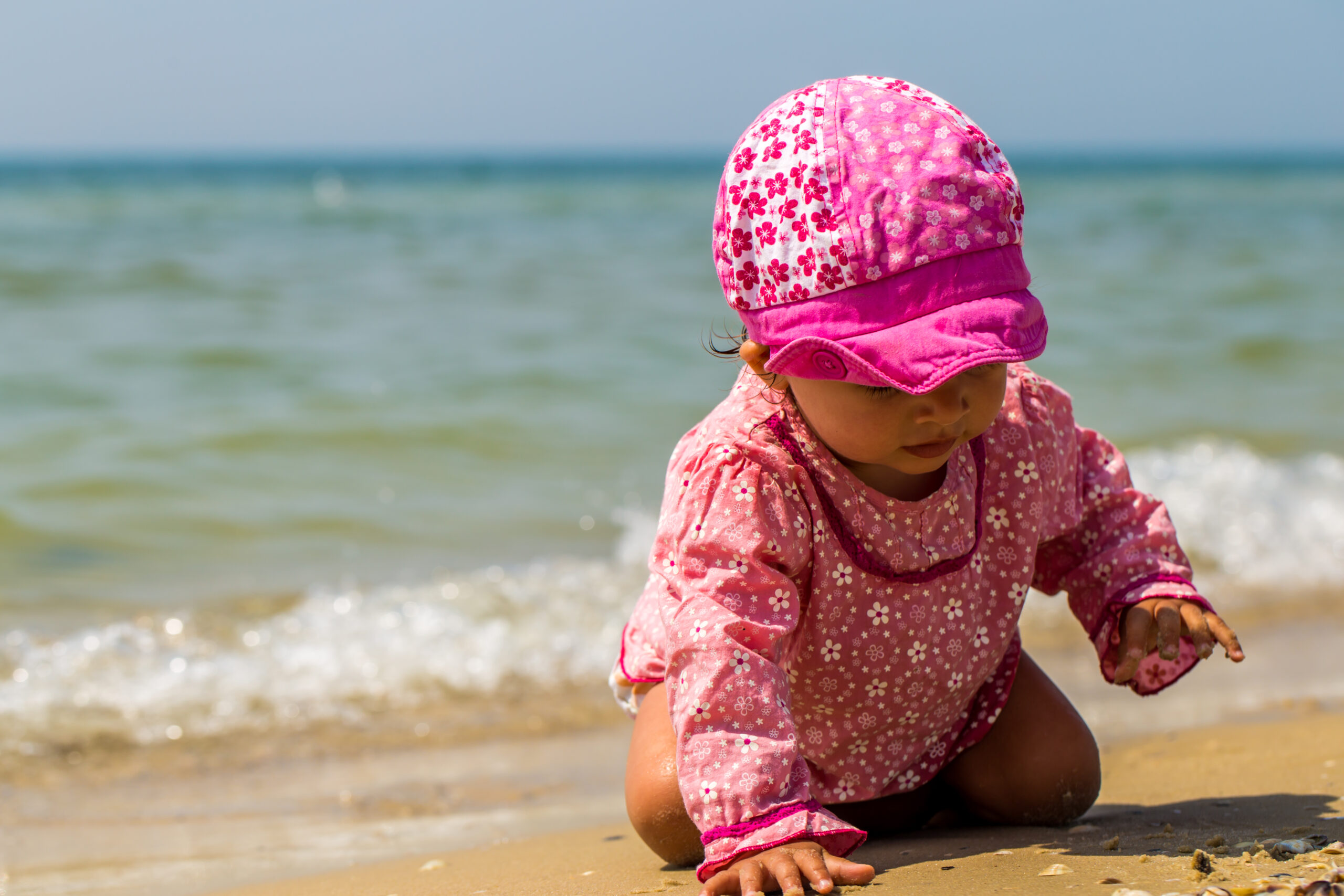 Blogfoto Zon op het strand: zo bescherm je je kind zonder gedoe Happy Kids Boutique zonbescherming met UV werende kleding voor kinderen op het strand waar een jong kind in roze outfit en petje speelt in het zand bij de zee met aandacht voor duurzaamheid
