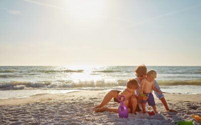 Ouder speelt met twee jonge kinderen in het zand op het strand bij zonsondergang met strand speelgoed, een warm familietaferel dat past bij Happy Kids Boutique waar babykleding en babybenodigdheden samenkomen met aandacht voor comfort en duurzaamheid.