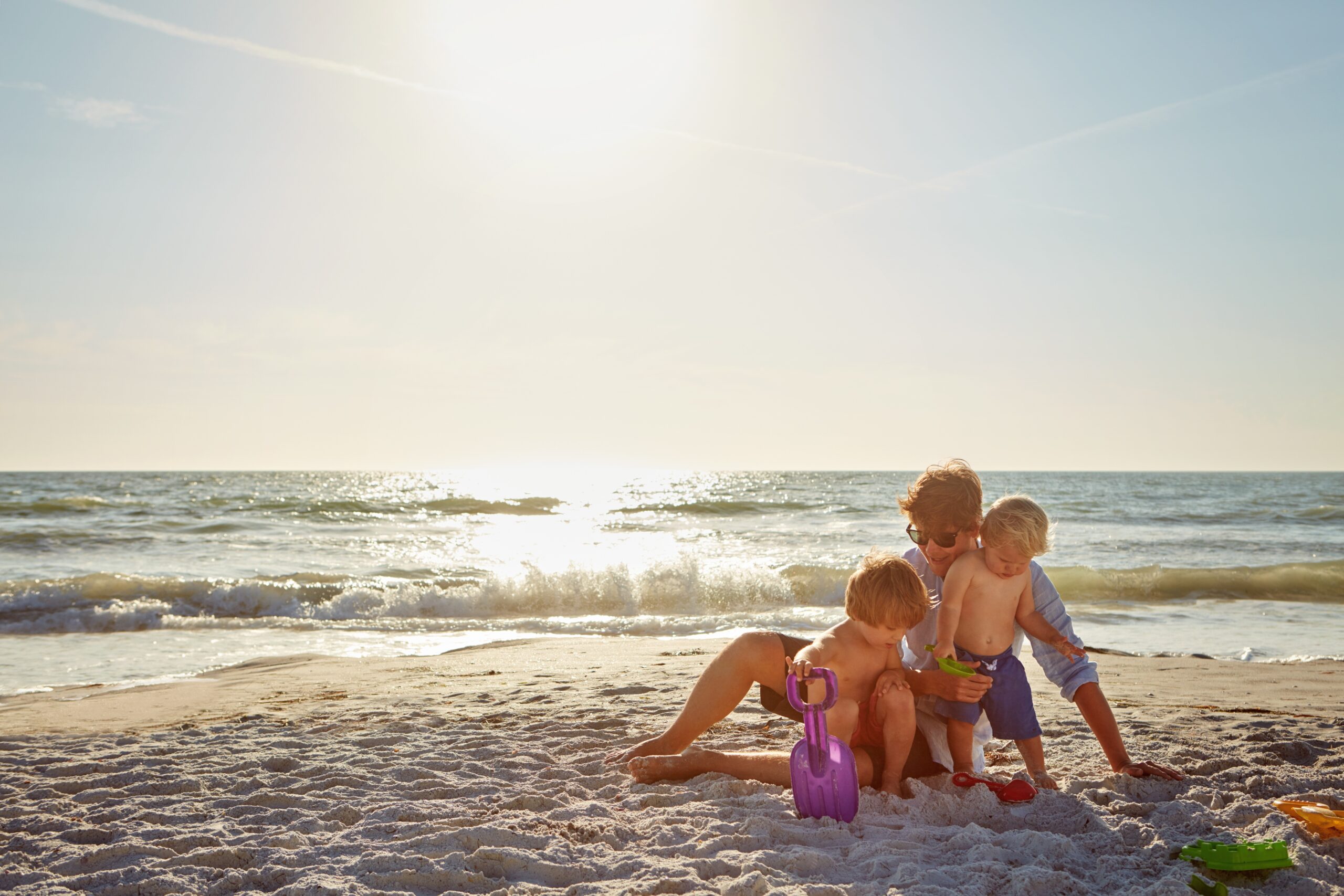 Ouder speelt met twee jonge kinderen in het zand op het strand bij zonsondergang met strand speelgoed, een warm familietaferel dat past bij Happy Kids Boutique waar babykleding en babybenodigdheden samenkomen met aandacht voor comfort en duurzaamheid.