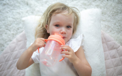 Happy Kids Boutique kind ligt ontspannen op een zacht kussen en is aan het drinken uit een drinkbeker Dr. Browns met handvatten, transparante beker met oranje deksel, focus op zelfstandig leren drinken en duurzaamheid in een rustige, lichte omgeving