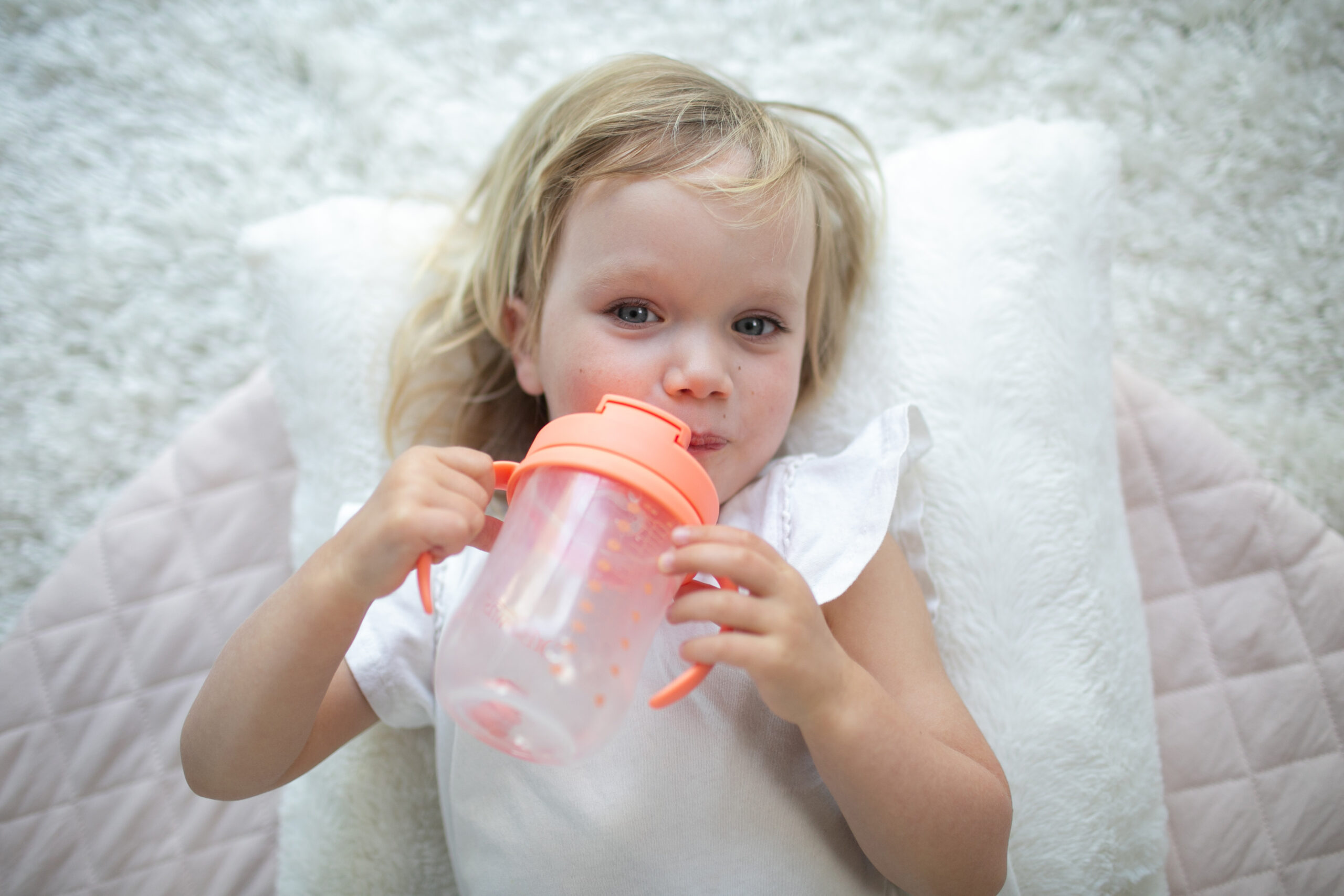 Happy Kids Boutique kind ligt ontspannen op een zacht kussen en is aan het drinken uit een drinkbeker Dr. Browns met handvatten, transparante beker met oranje deksel, focus op zelfstandig leren drinken en duurzaamheid in een rustige, lichte omgeving