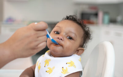 Happy Kids Boutique afbeelding van een baby in een kinderstoel die eerste hapjes krijgt met een lepel, waarbij een ouder voorzichtig voeding aanbiedt met een Dr. Brown's lepel in een rustige keukenomgeving