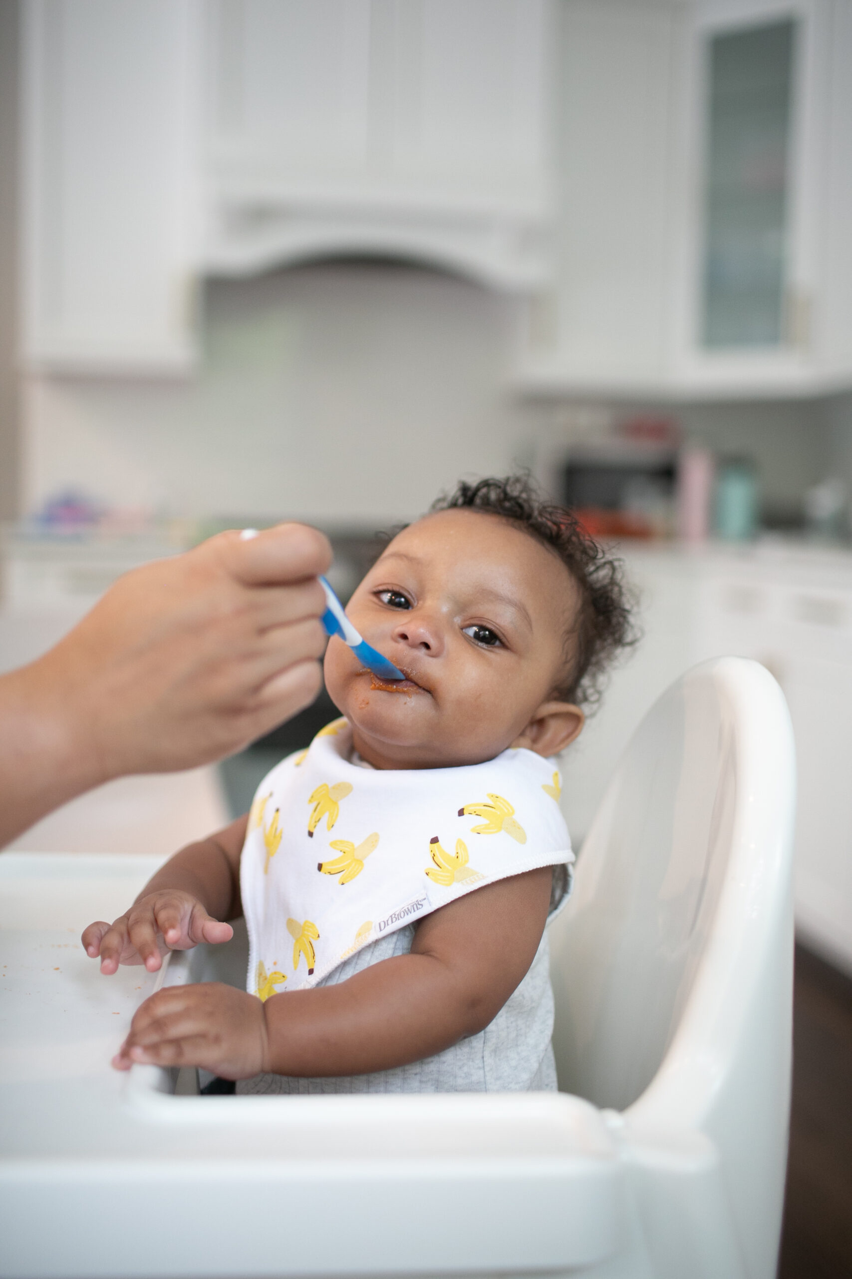 Happy Kids Boutique afbeelding van een baby in een kinderstoel die eerste hapjes krijgt met een lepel, waarbij een ouder voorzichtig voeding aanbiedt met een Dr. Brown's lepel in een rustige keukenomgeving