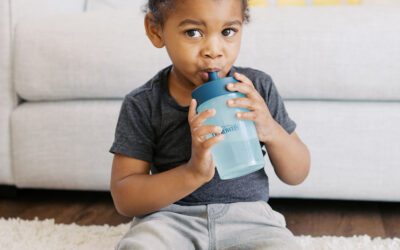 Happy Kids Boutique kind dat op de grond zit en zelfstandig drinkt uit een Dr. Brown beker met rietje, ontspannen moment in huiselijke sfeer met focus op drinken en ontwikkeling