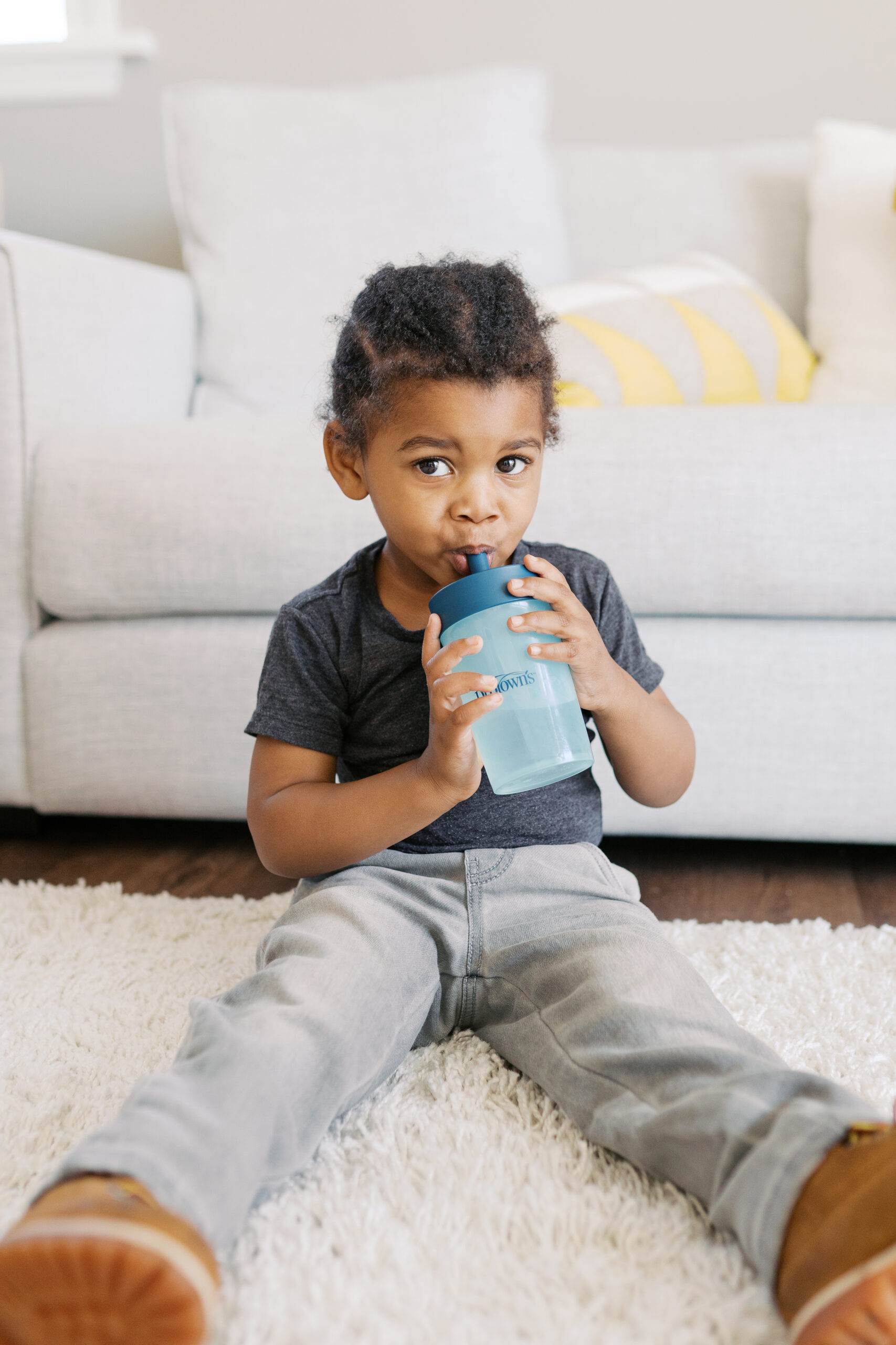 Happy Kids Boutique kind dat op de grond zit en zelfstandig drinkt uit een Dr. Brown beker met rietje, ontspannen moment in huiselijke sfeer met focus op drinken en ontwikkeling