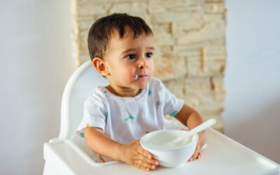 Happy Kids Boutique kind wil niet eten dat in een kinderstoel zit met een kommetje en lepel in de hand, twijfelend en afgeleid tijdens het eetmoment in een rustige huiselijke omgeving met aandacht voor duurzaamheid