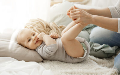 Happy Kids Boutique baby ligt ontspannen op een zacht kleed terwijl ouder de beentjes vasthoudt, rustige lifestyle afbeelding bij babymassage en het liefdevol masseren van je baby