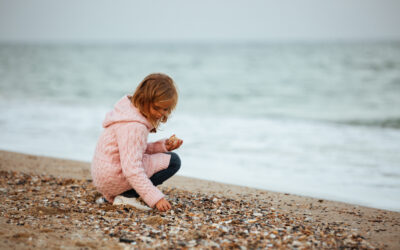 Happy Kids Boutique kind hurkt op het strand tussen schelpen en zoekt aandachtig naar mooie vondsten langs de waterlijn, zacht gekleed in een roze vest, spelend en ontdekkend met oog voor natuur en duurzaamheid