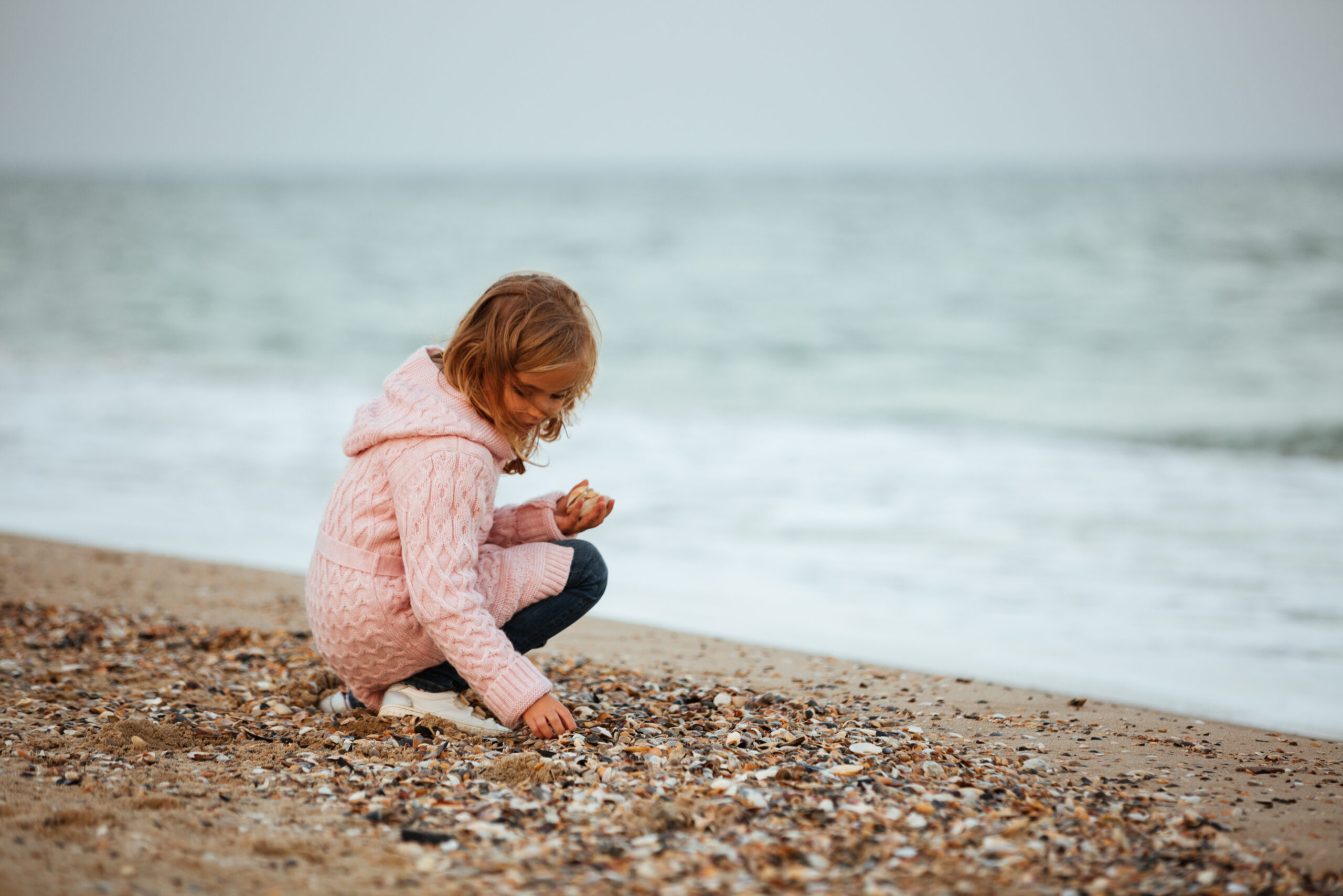 Happy Kids Boutique kind hurkt op het strand tussen schelpen en zoekt aandachtig naar mooie vondsten langs de waterlijn, zacht gekleed in een roze vest, spelend en ontdekkend met oog voor natuur en duurzaamheid