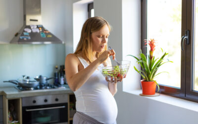 Happy Kids Boutique zwangere vrouw staat in de keuken bij het raam en is aan het eten van een verse salade uit een glazen kom, rustige en lichte setting die het moment van zwangerschap en bewust eten benadrukt