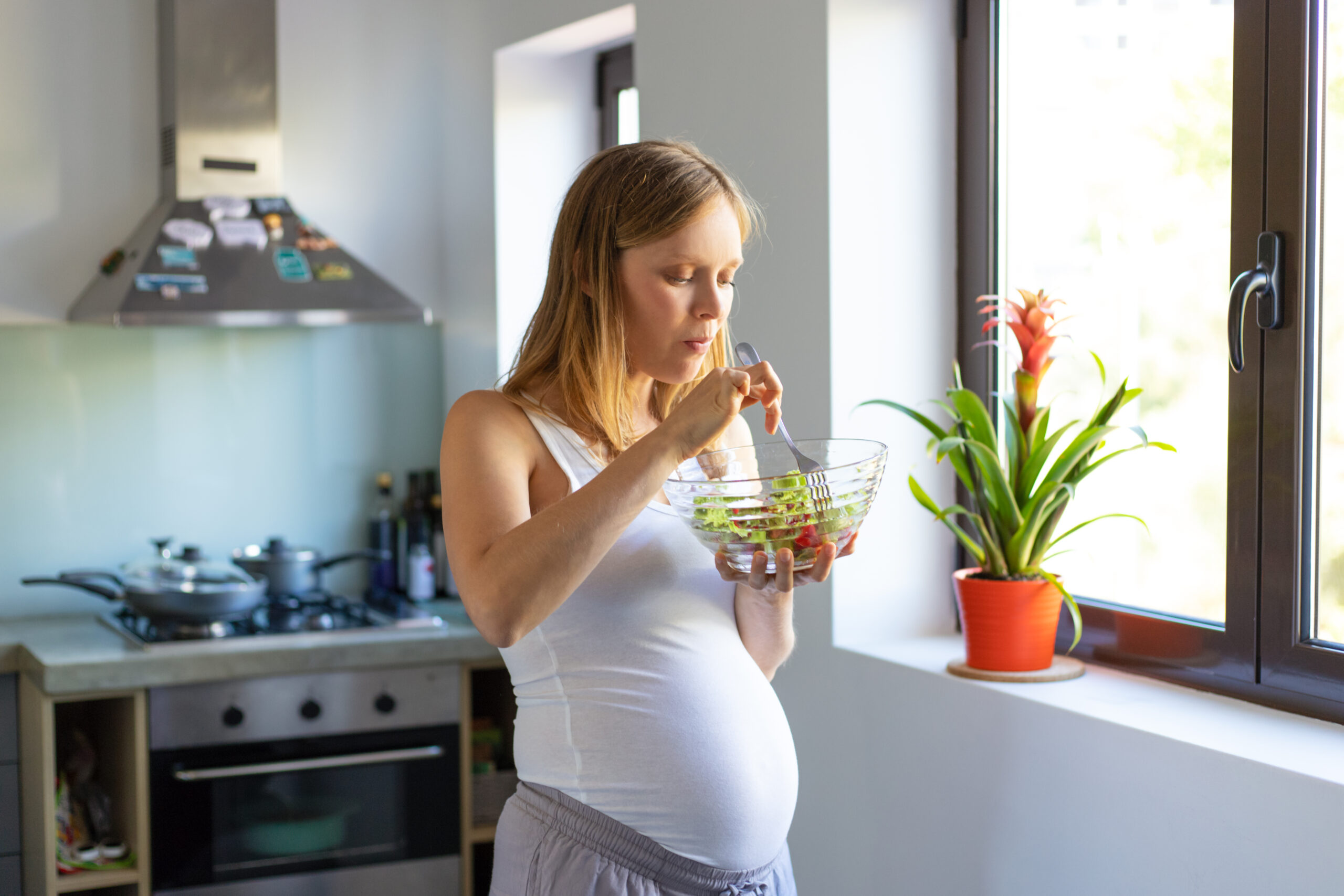 Happy Kids Boutique zwangere vrouw staat in de keuken bij het raam en is aan het eten van een verse salade uit een glazen kom, rustige en lichte setting die het moment van zwangerschap en bewust eten benadrukt