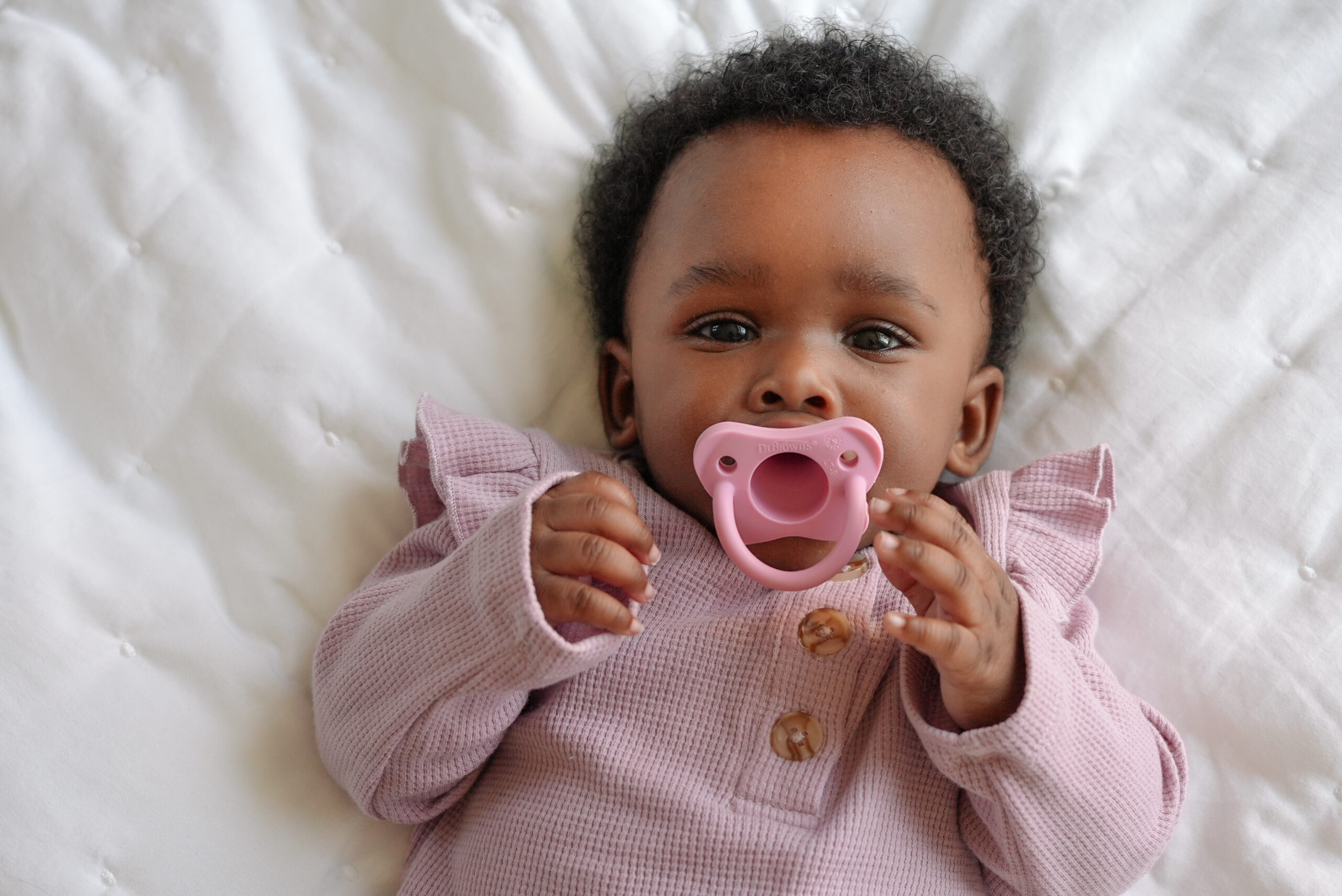 Happy Kids Boutique baby met roze Dr. Brown's fopspeen liggend op een wit dekentje, kindje draagt roze kleding en kijkt rustig naar de camera, lifestyle afbeelding van een comfortabele fopspeen voor dagelijks gebruik