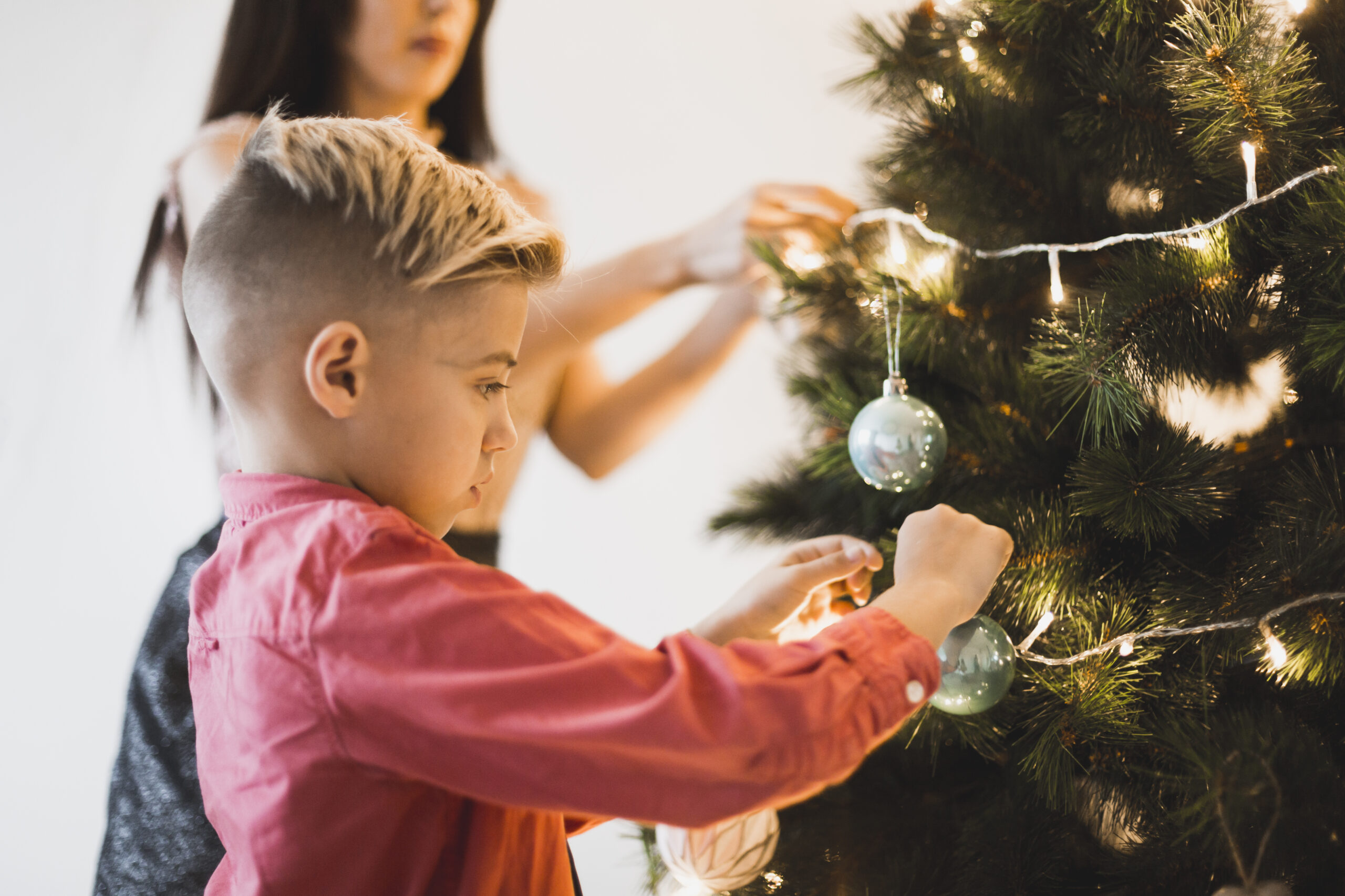 Blogfoto kerst en geloof Happy Kids Boutique kind versiert samen met volwassene de kerstboom met lichtjes en kerstballen, warme lifestyle afbeelding over kerst, geloof en samen momenten beleven tijdens de feestdagen