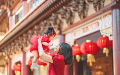 Happy Kids Boutique moeder tilt haar baby in de lucht tijdens Chinees Nieuwjaar, beiden gekleed in rood voor de viering van culturen, met traditionele lantaarns en versierde tempel op de achtergrond, een warm en duurzaam familiemoment