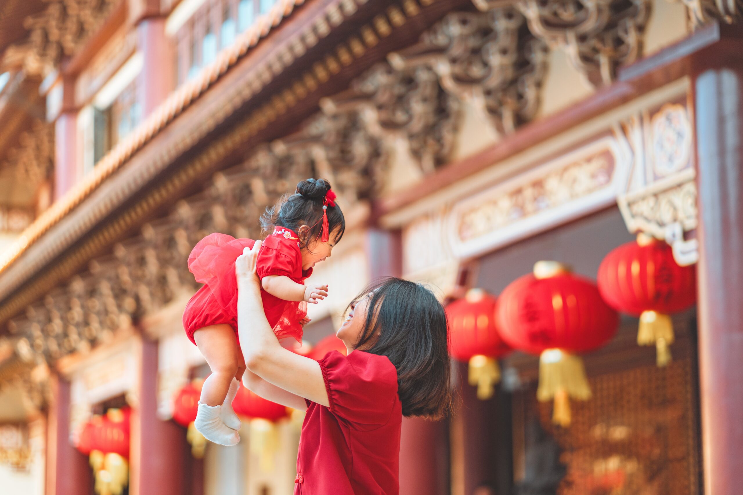 Happy Kids Boutique moeder tilt haar baby in de lucht tijdens Chinees Nieuwjaar, beiden gekleed in rood voor de viering van culturen, met traditionele lantaarns en versierde tempel op de achtergrond, een warm en duurzaam familiemoment