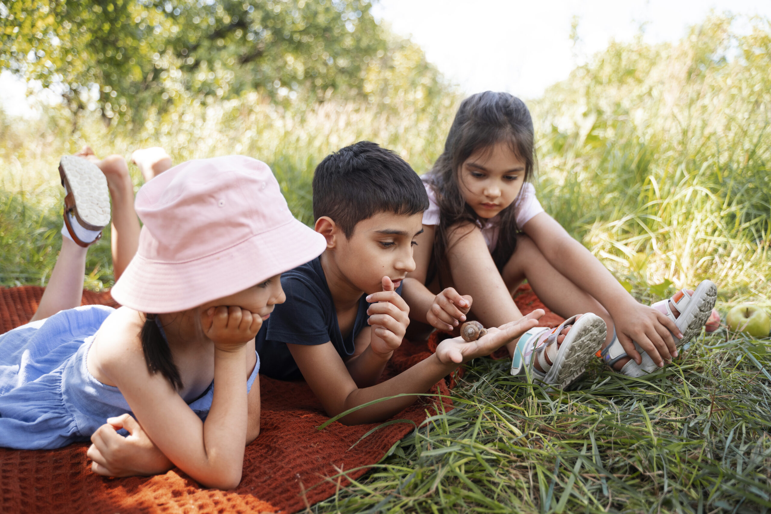 Blogfoto kennismaken met de wereld Happy Kids Boutique kinderen liggen samen op een kleed in de natuur en bekijken een slak, vrolijke lifestyle afbeelding over vakantie, nieuwe ontdekkingen en kennismaken met de wereld.