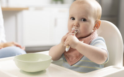 Baby in een kinderstoel eet zelfstandig de eerste vaste voeding met een lepeltje.