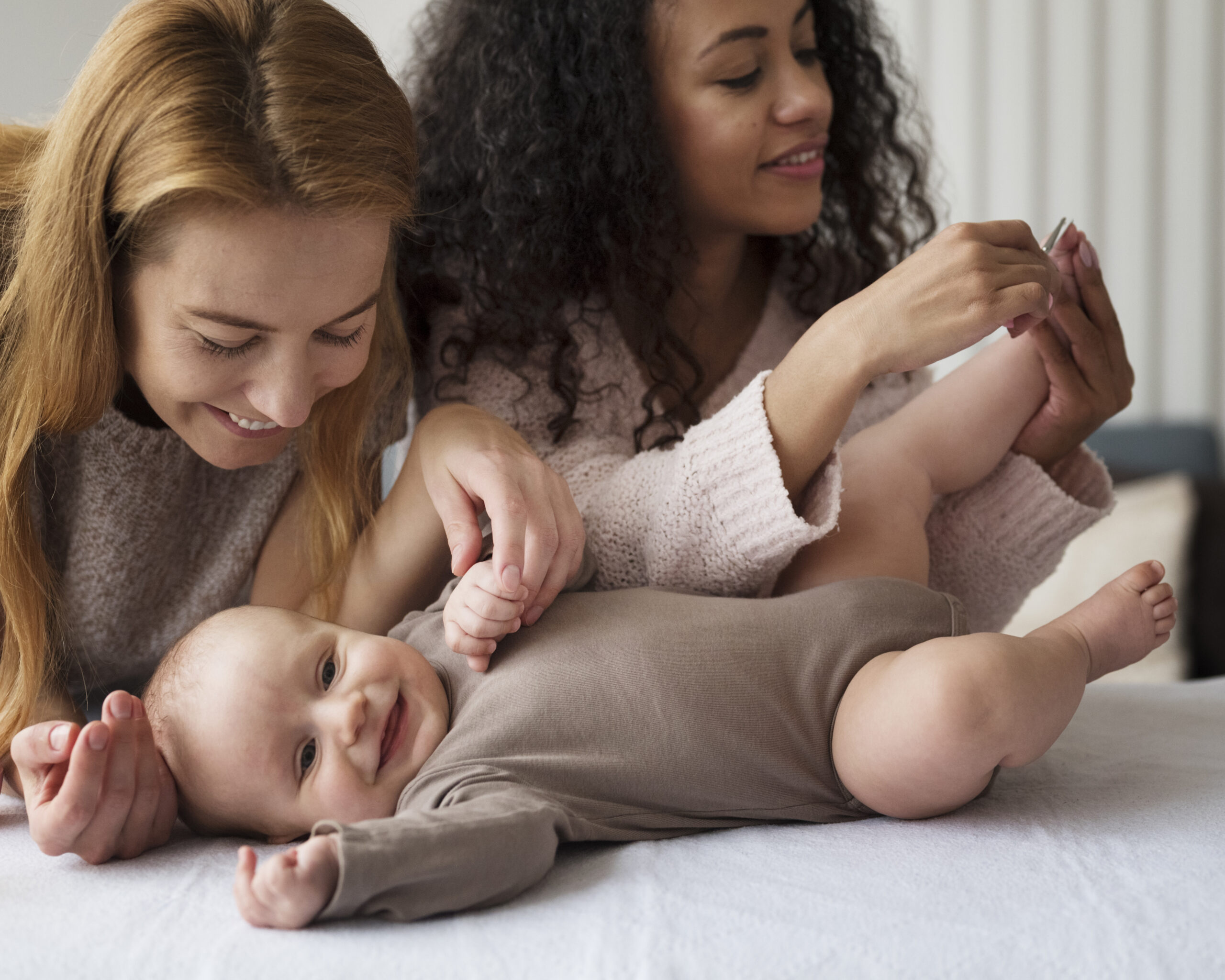 Twee moeders verzorgen samen een lachende baby tijdens een rustig verzorgingsmoment thuis.