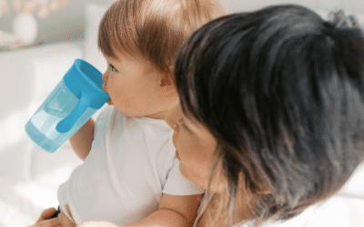 Kindje drinkt zelfstandig uit een blauwe meegroeiende drinkbeker terwijl het bij een ouder op schoot zit.