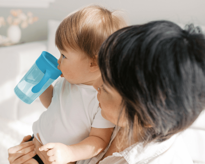 Kindje drinkt zelfstandig uit een blauwe meegroeiende drinkbeker terwijl het bij een ouder op schoot zit.
