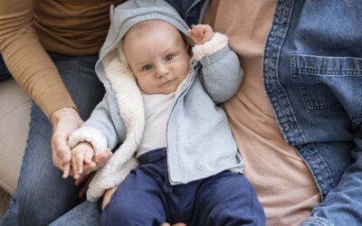 Baby in een warm vestje zit op schoot tussen twee ouders en kijkt nieuwsgierig naar de camera.