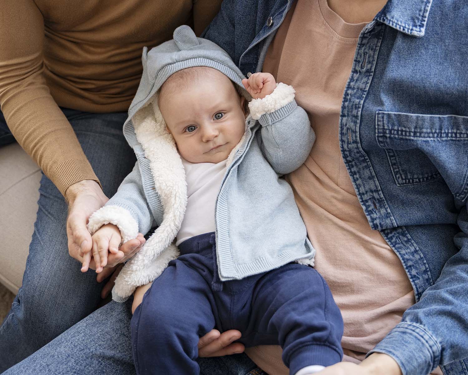Baby in een warm vestje zit op schoot tussen twee ouders en kijkt nieuwsgierig naar de camera.