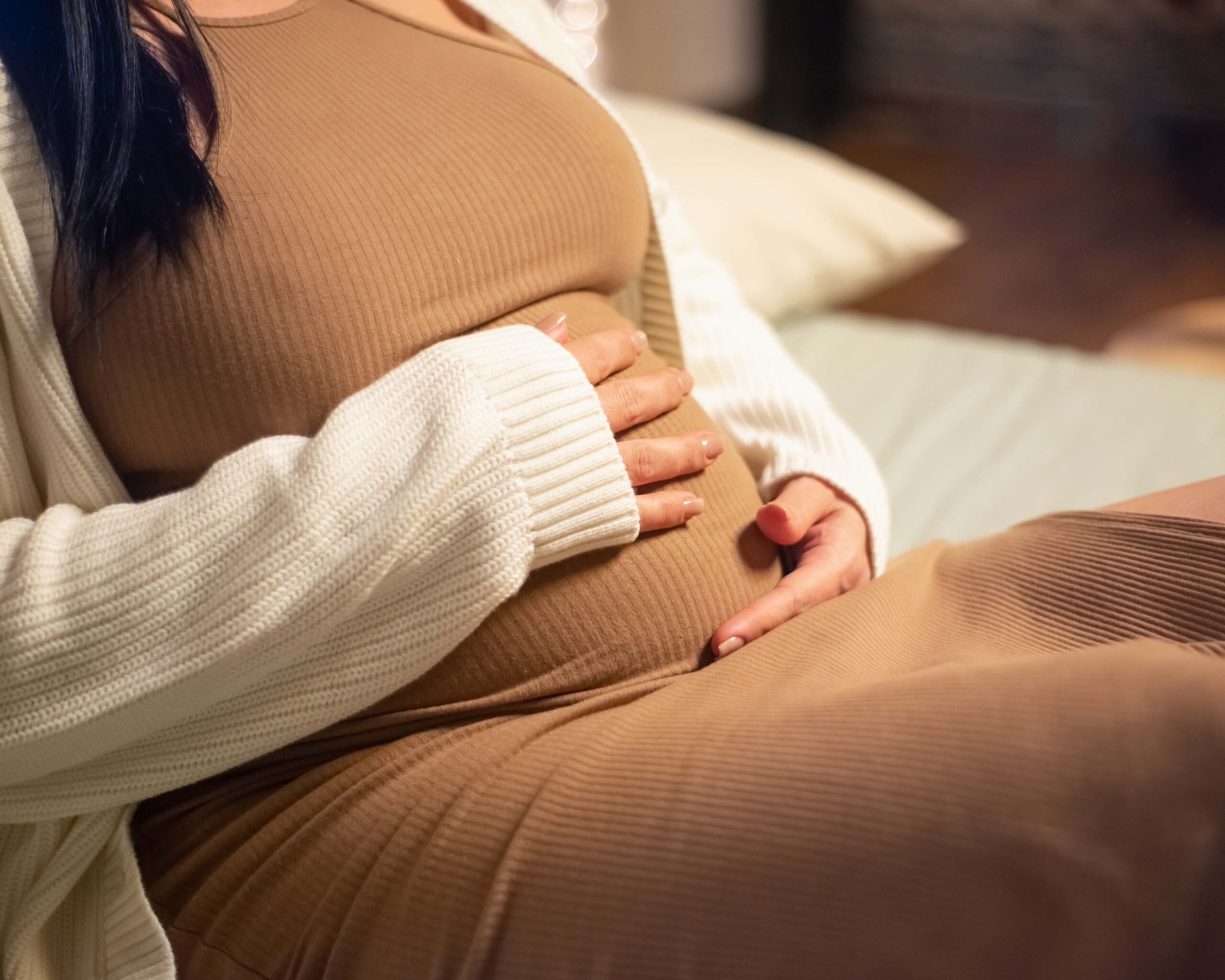 Zwangere vrouw zit ontspannen op de bank en houdt haar handen liefdevol op haar babybuik tijdens de voorbereiding op de komst van haar kleintje.