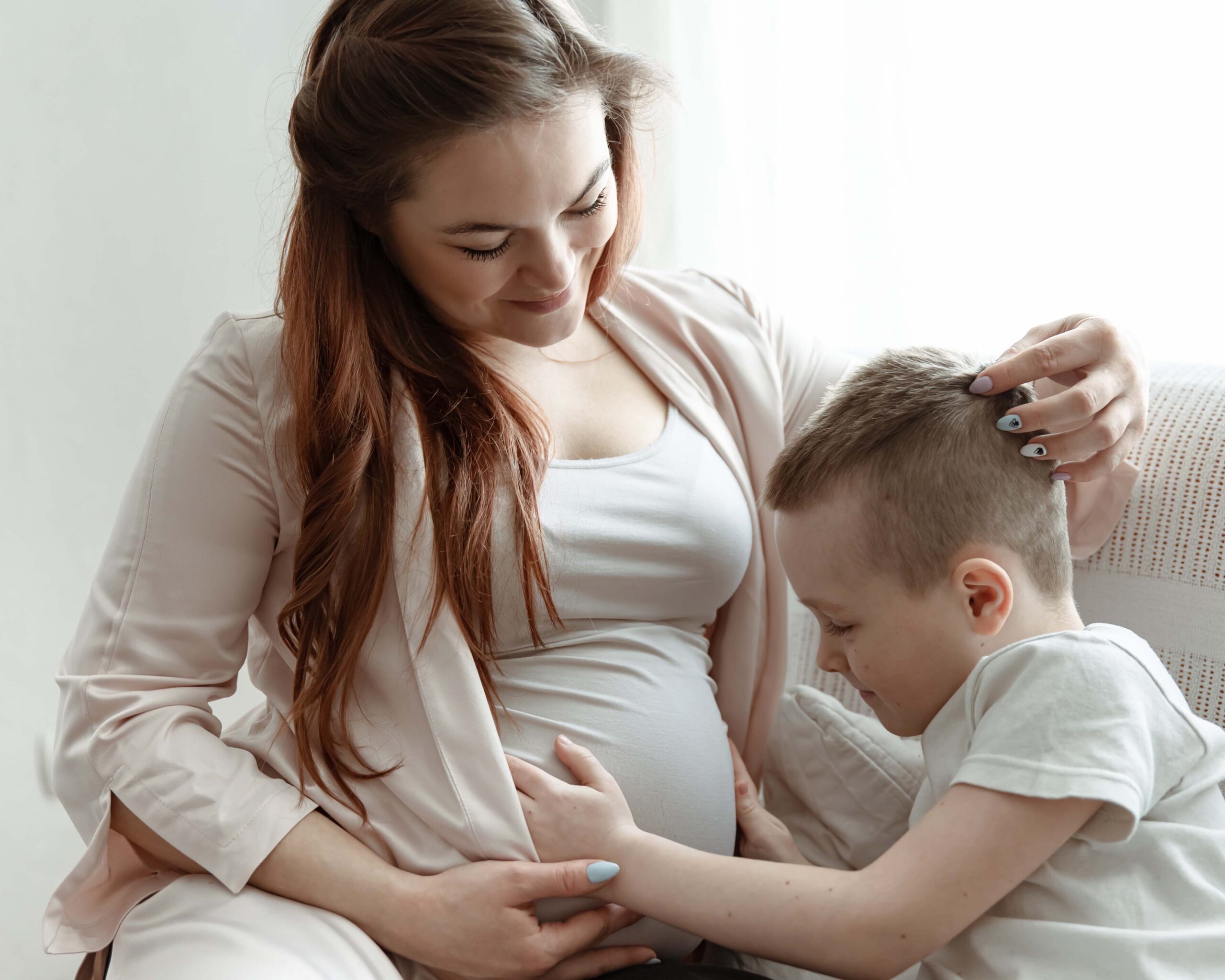 Zwangere moeder zit samen met haar zoontje op de bank, terwijl hij liefdevol haar babybuik aanraakt in afwachting van zijn broertje of zusje.